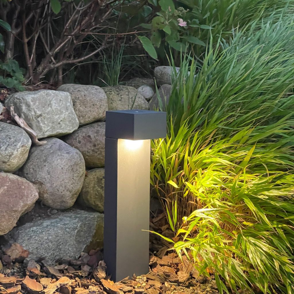 Eine moderne Gartenlampe, die sanftes Licht abgibt, steht neben einem Steinmauer und grünen Pflanzen.