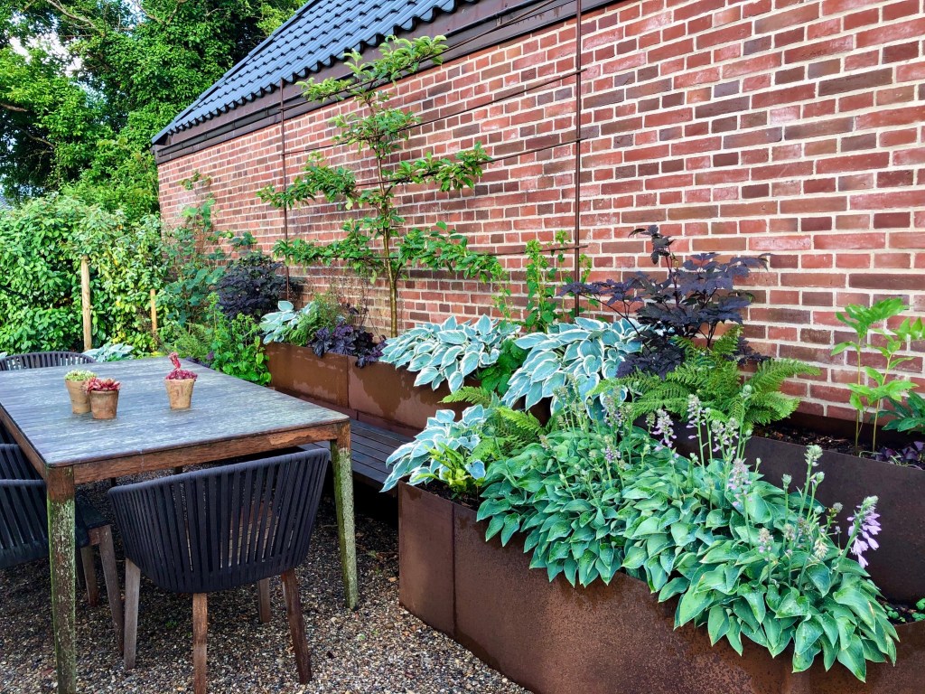 Ein stilvoller Gartenbereich mit einem Holztisch und Stühlen. Im Hintergrund sind verschiedene Pflanzen in modernen Pflanzkästen angeordnet, vor einer Backsteinmauer.