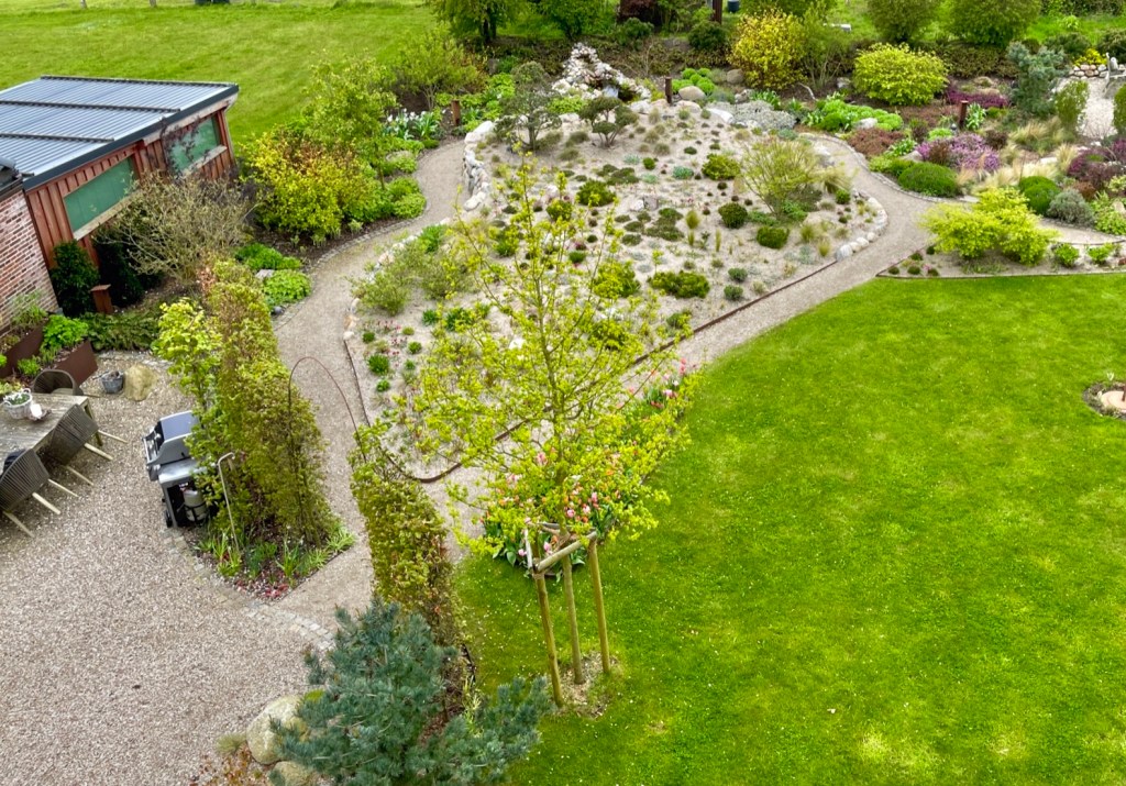 Luftaufnahme eines gepflegten Gartens mit einem Kiesweg, Blumenbeeten und einer Rasenfläche. Im Hintergrund ist ein Holzschuppen sichtbar.
