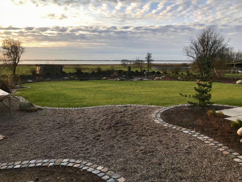 Blick auf einen gepflegten Garten mit grünem Rasen, umgeben von Kieswegen und Bäumen, bei Sonnenuntergang.