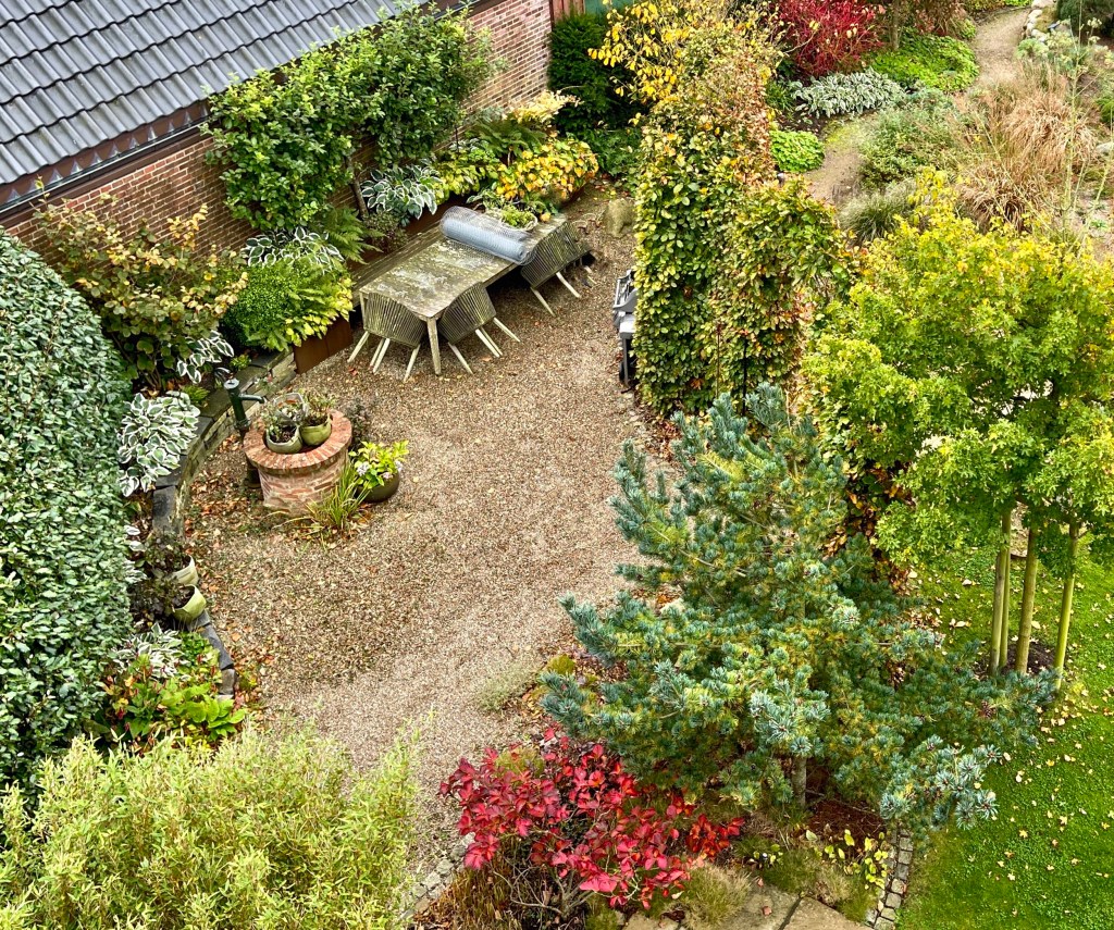 Eine Luftaufnahme eines Gartens mit bunten Sträuchern, Bäumen und einem Steingarten. In der Mitte steht ein Tisch mit Stühlen, umgeben von Pflanzen in verschiedenen Farben.